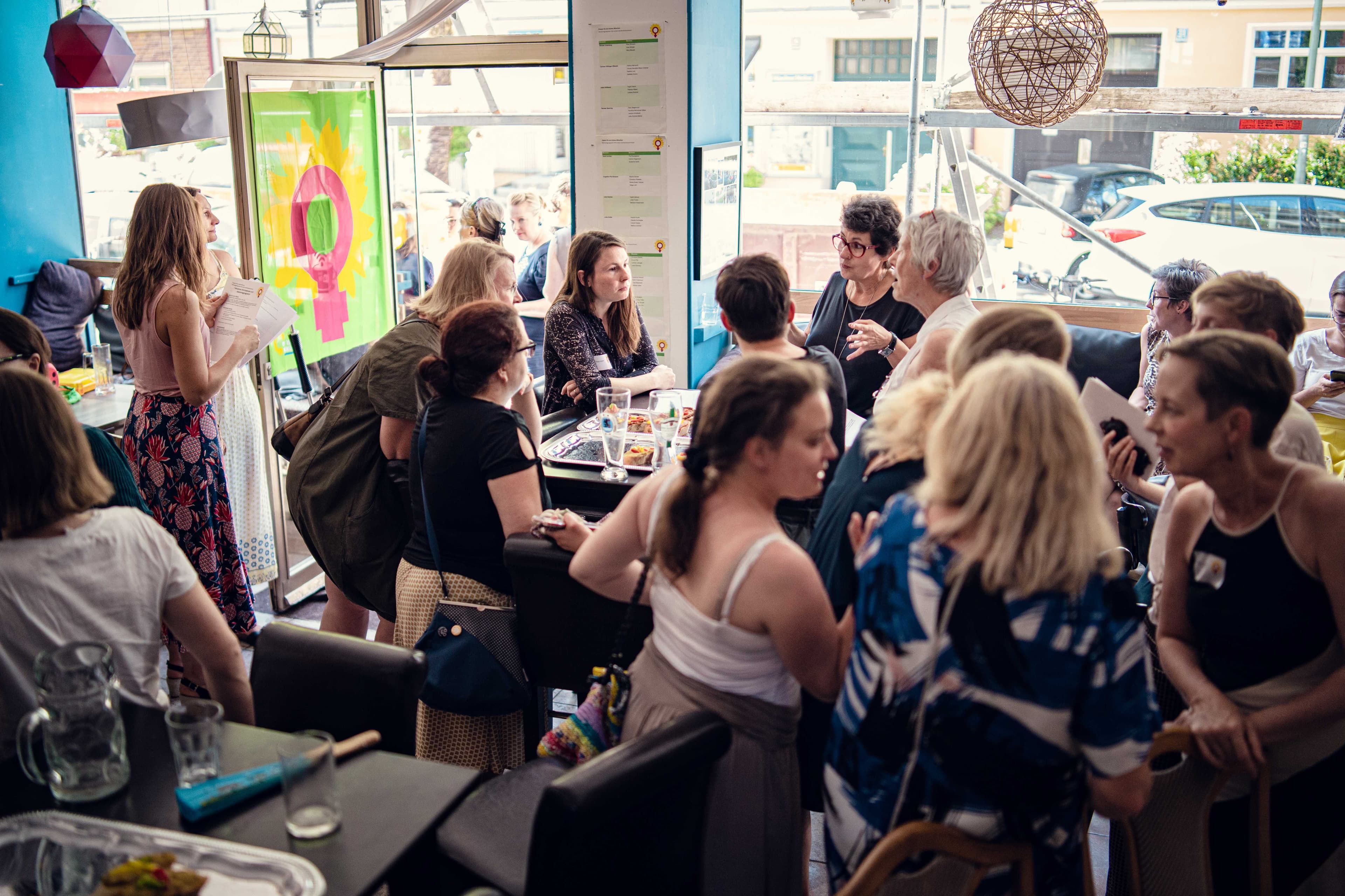 Viele Frauen diskutieren in einem Raum. Im Hintergrund ist die grüne Flagge mit dem Frauensymbol zu sehen.